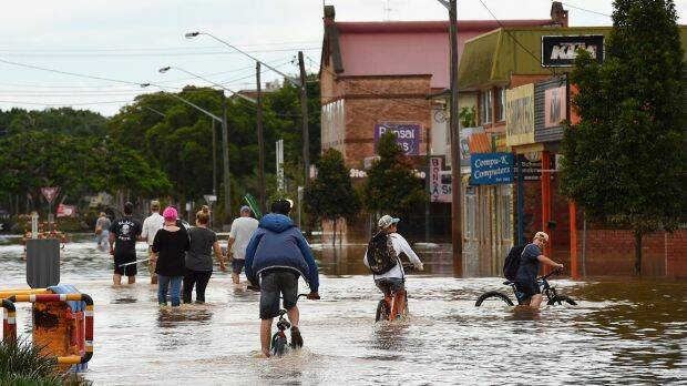 Teenagers cycle behind a group of people with brooms wading through the water of the flooded streets of Lismore. Photo: Kate Geraghty
